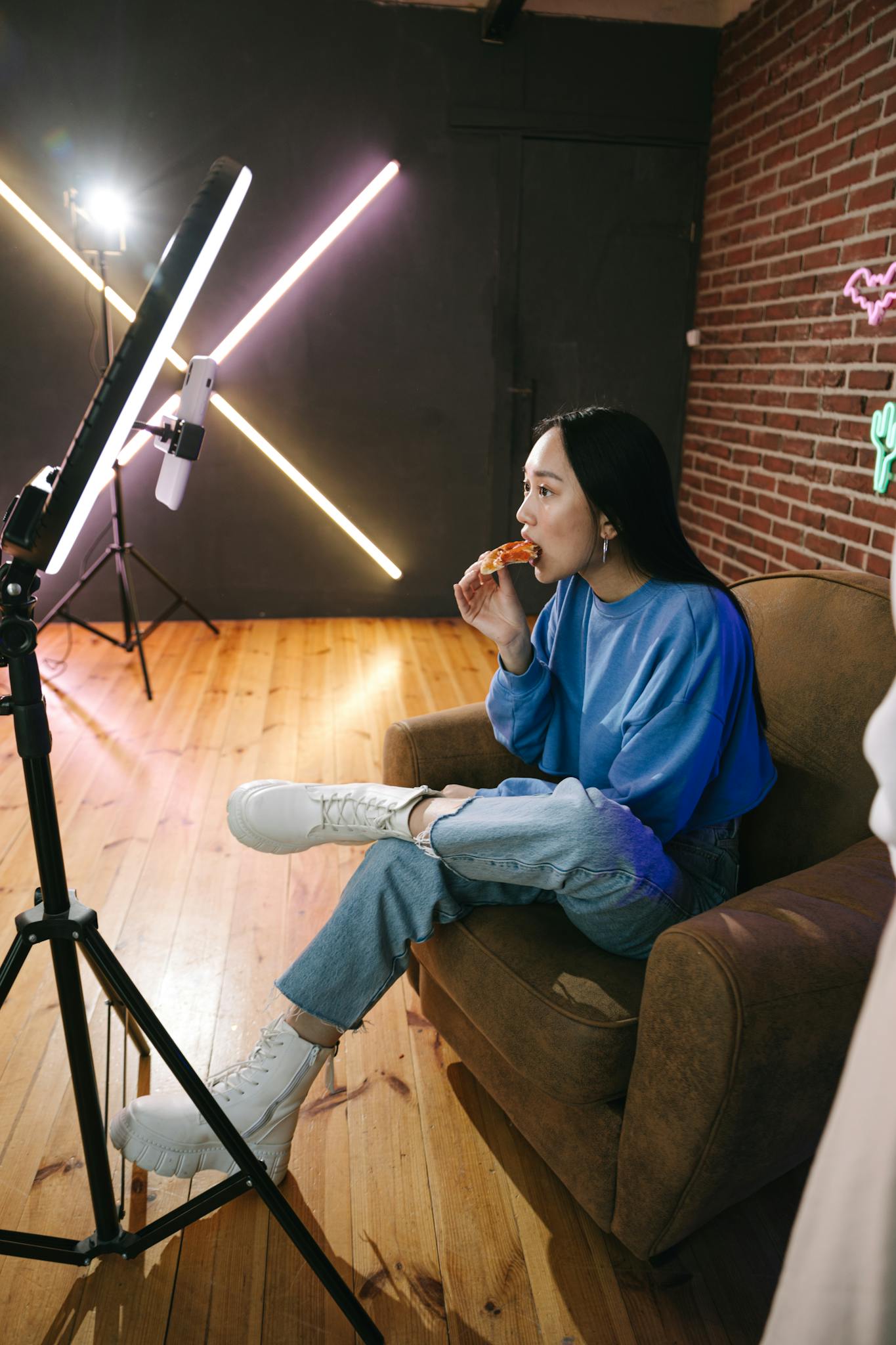 A young woman eats pizza while streaming in a stylish indoor setup with neon lights.