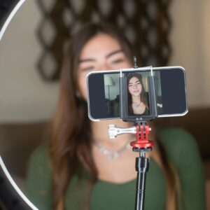 A young woman live streams from home using a smartphone and ring light setup.